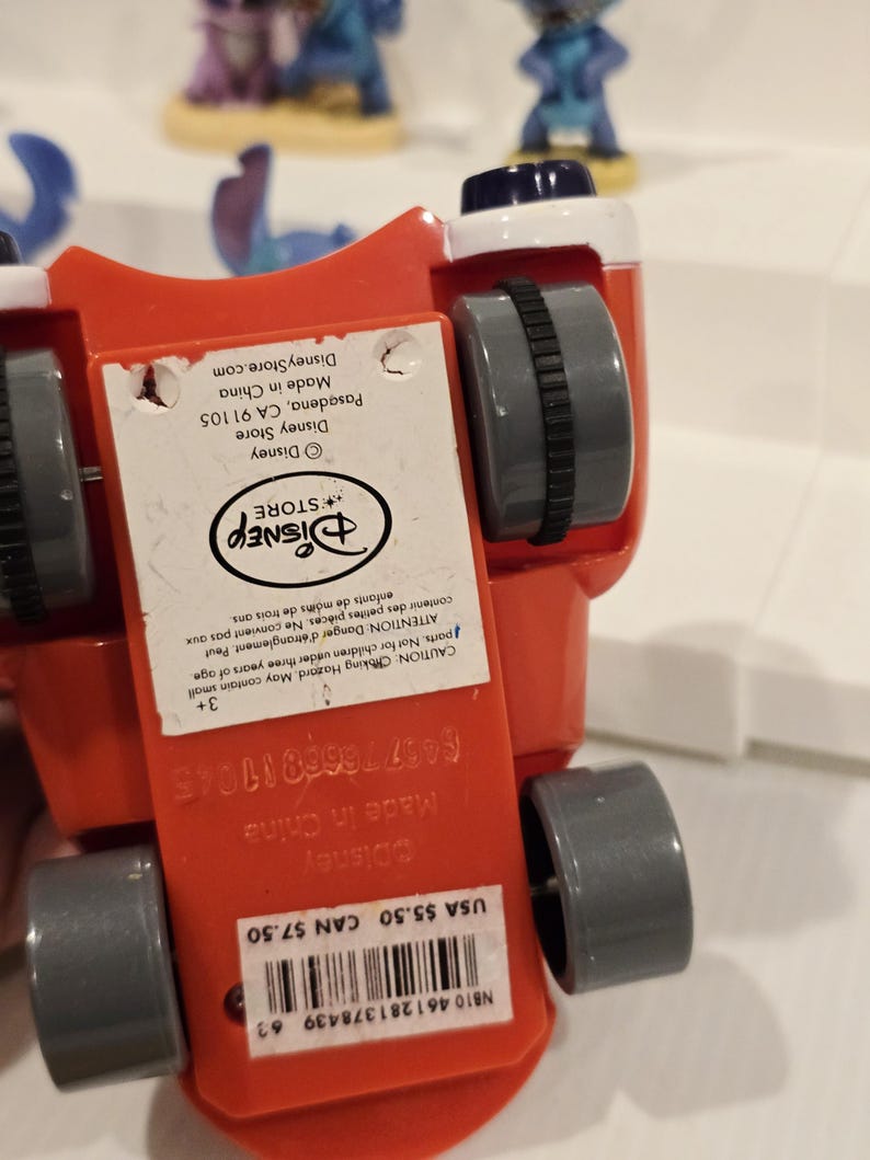 May include: A red toy car with gray wheels and a white and black top. The underside of the car has a Disney Store label with text. Other Stitch figurines are visible in the background.