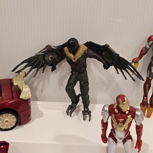 May include: A collection of action figures featuring characters from a superhero franchise. The Vulture figure has large, articulated wings. Other figures include Iron Man and a red car toy. The figures are on a white surface.