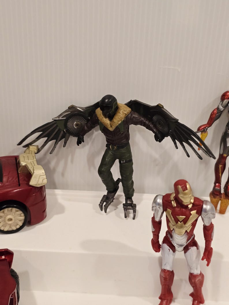 May include: A collection of action figures featuring characters from a superhero franchise. The Vulture figure has large, articulated wings. Other figures include Iron Man and a red car toy. The figures are on a white surface.