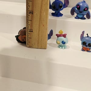 May include: A collection of small, colorful Stitch figurines, each with unique poses and expressions. The figurines are blue, with some featuring pink accents. A wooden ruler, approximately 3 inches long, is in the image for scale.