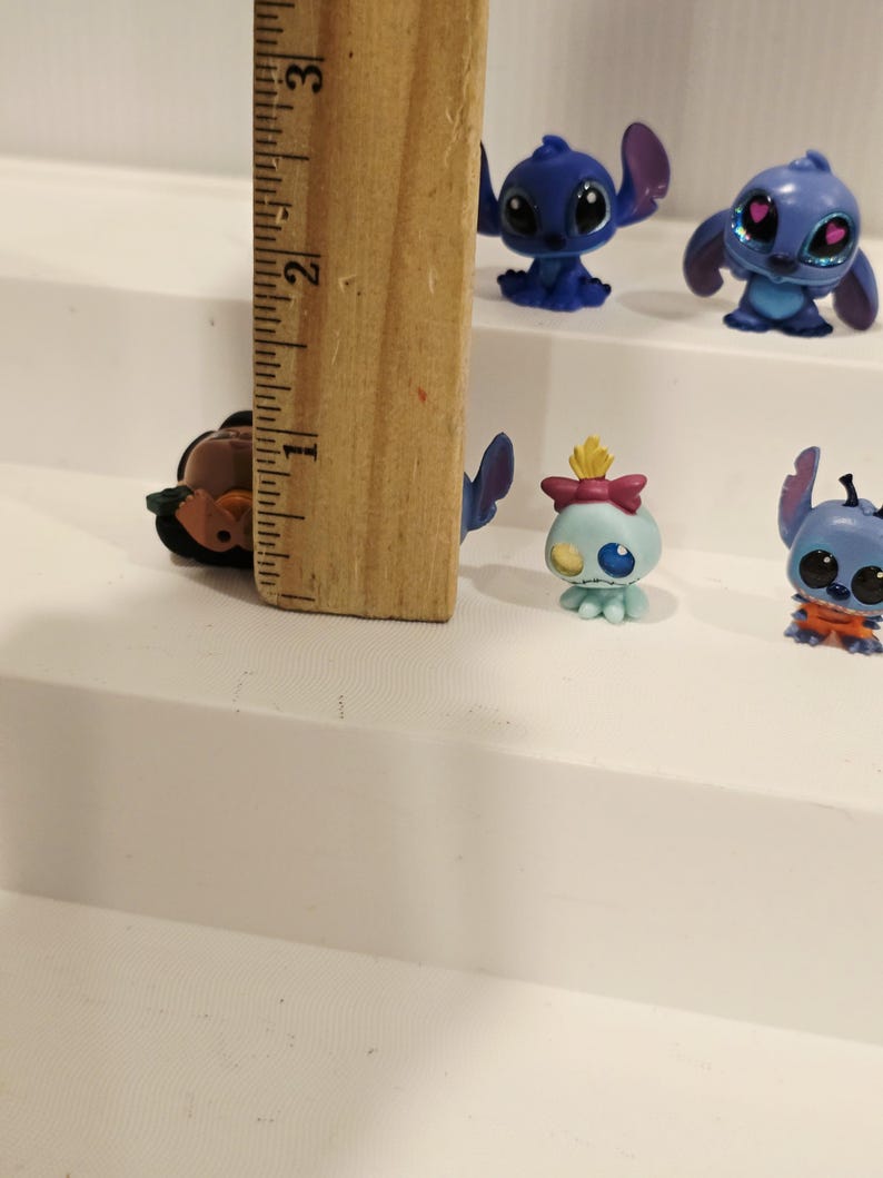May include: A collection of small, colorful Stitch figurines, each with unique poses and expressions. The figurines are blue, with some featuring pink accents. A wooden ruler, approximately 3 inches long, is in the image for scale.