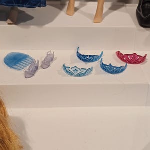 May include: A selection of doll accessories, including tiaras, shoes, and a comb. The tiaras are in shades of blue and pink. The shoes and comb are a translucent purple. The items are arranged on a white surface.