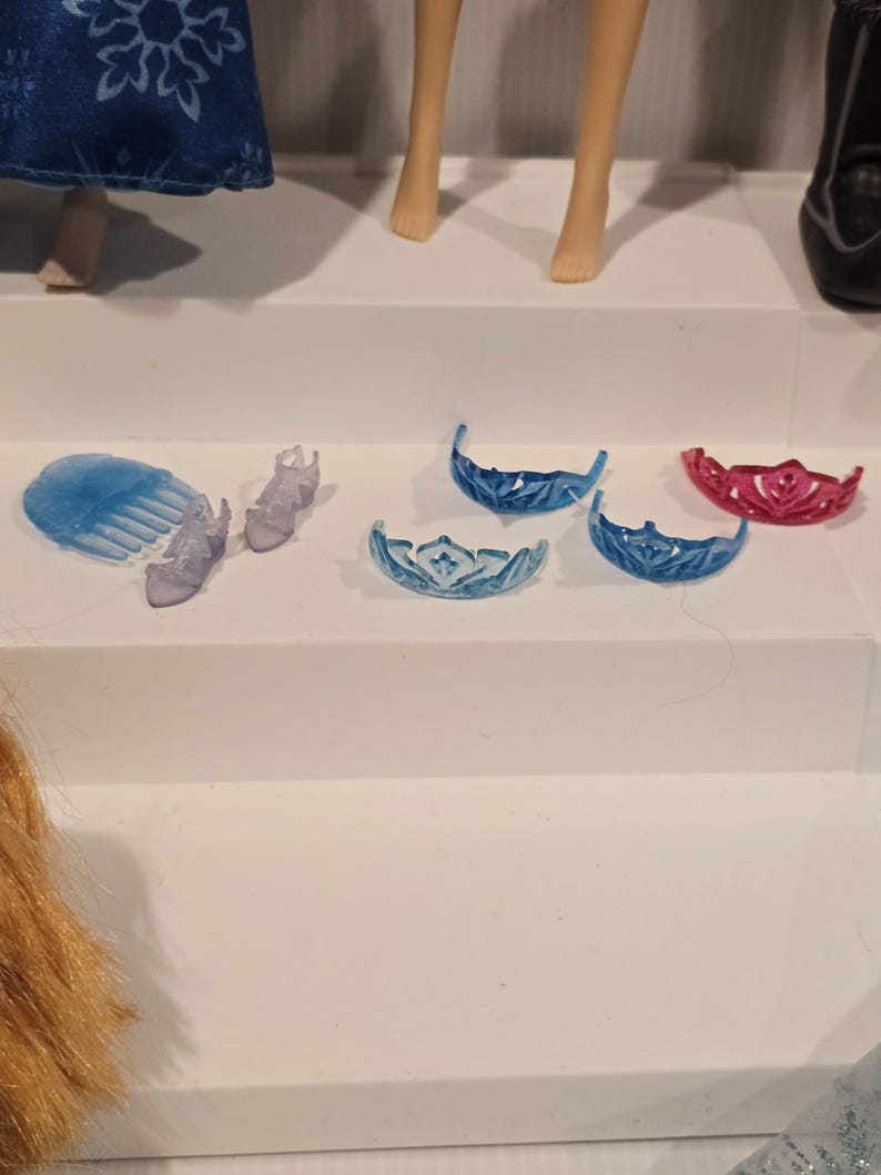 May include: A selection of doll accessories, including tiaras, shoes, and a comb. The tiaras are in shades of blue and pink. The shoes and comb are a translucent purple. The items are arranged on a white surface.