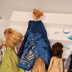 May include: A collection of dolls, one with a blue cape and gold detailing, another in a green dress. Accessories are placed on a white shelf. The dolls have different hair colours and styles. A children's toy.