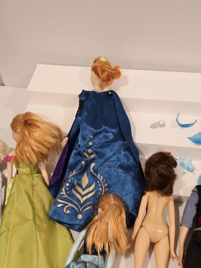 May include: A collection of dolls, one with a blue cape and gold detailing, another in a green dress. Accessories are placed on a white shelf. The dolls have different hair colours and styles. A children's toy.