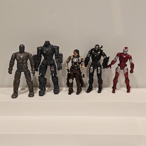 May include: Five action figures of Iron Man characters in various armor designs. The figures are in shades of gray, black, brown, and red. The figures are standing on a white surface.