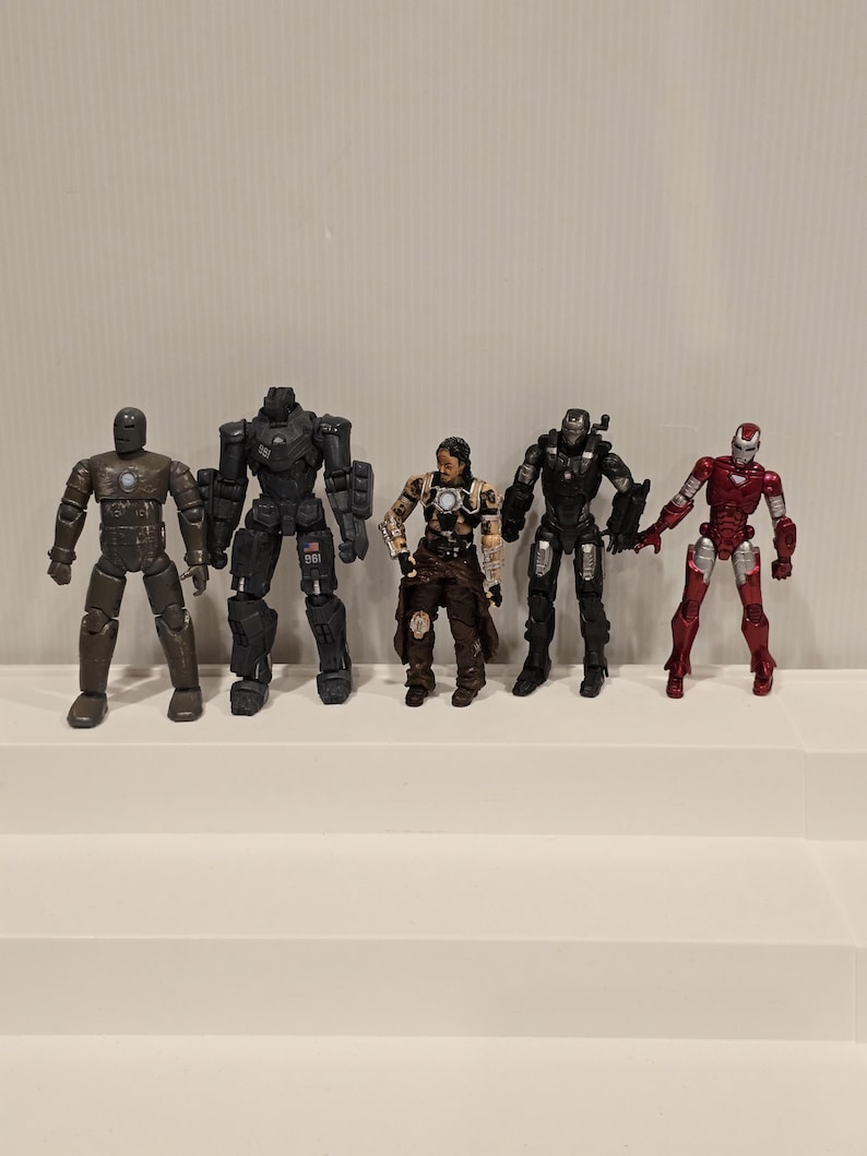 May include: Five action figures of Iron Man characters in various armor designs. The figures are in shades of gray, black, brown, and red. The figures are standing on a white surface.