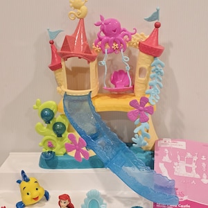May include: A colorful plastic playset featuring a castle with a slide, a swing, and various sea creature figurines. The castle has pink and yellow accents, with a blue slide. The figurines include a yellow fish, a red crab, and a doll.