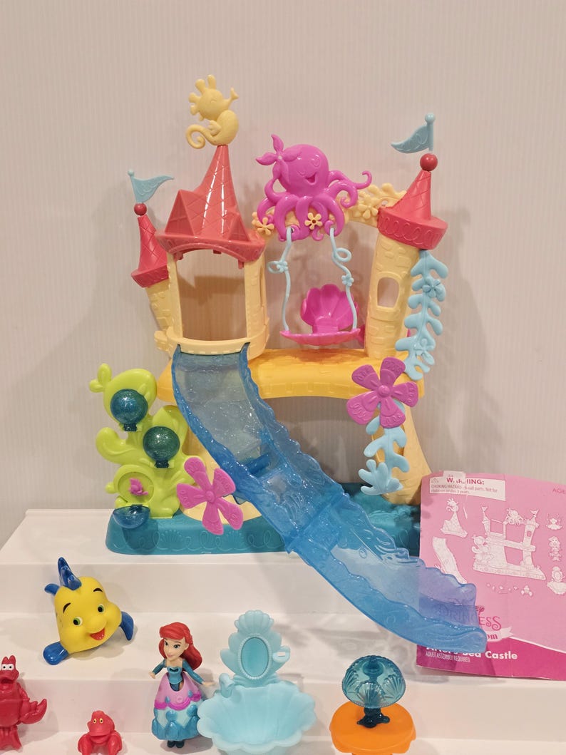 May include: A colorful plastic playset featuring a castle with a slide, a swing, and various sea creature figurines. The castle has pink and yellow accents, with a blue slide. The figurines include a yellow fish, a red crab, and a doll.