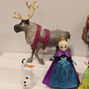 May include: A collection of Disney Frozen character figurines. Includes a gray reindeer with cream antlers and pink harness, a white snowman with a carrot nose, and a doll in a blue dress and purple cape.