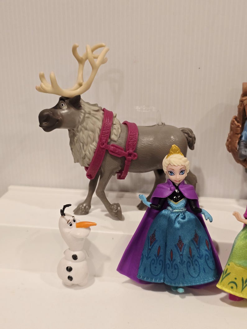 May include: A collection of Disney Frozen character figurines. Includes a gray reindeer with cream antlers and pink harness, a white snowman with a carrot nose, and a doll in a blue dress and purple cape.