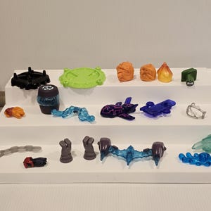 May include: A collection of colorful plastic toy parts, including a black base, a green disc, orange and yellow flame-shaped pieces, and various blue and purple components. The items are arranged on a tiered white display.