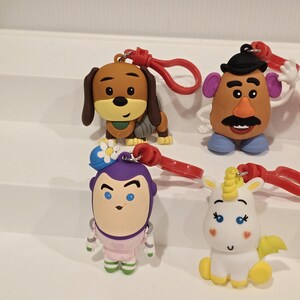 May include: A selection of character keyrings, including a brown dog, Mr. Potato Head, Buzz Lightyear, and a white unicorn. Each keyring has a red clasp. The characters are brightly coloured and detailed, perfect for attaching to keys or bags.
