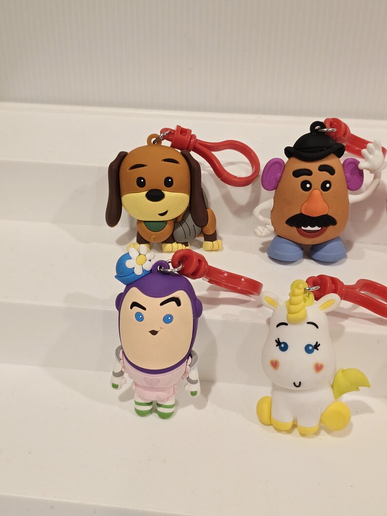 May include: A selection of character keyrings, including a brown dog, Mr. Potato Head, Buzz Lightyear, and a white unicorn. Each keyring has a red clasp. The characters are brightly coloured and detailed, perfect for attaching to keys or bags.