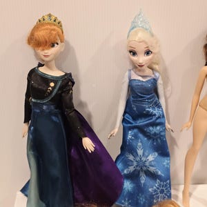 May include: Two dolls of animated characters, one with red hair and a gold crown, wearing a black and blue dress with a purple skirt. The other doll has blonde hair and a blue dress with a snowflake pattern. Additional doll heads are visible.