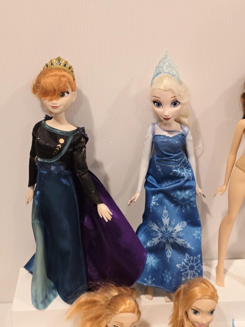May include: Two dolls of animated characters, one with red hair and a gold crown, wearing a black and blue dress with a purple skirt. The other doll has blonde hair and a blue dress with a snowflake pattern. Additional doll heads are visible.
