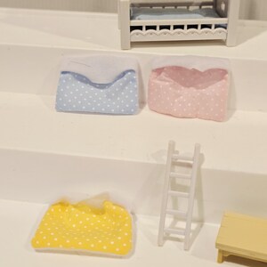 Puede incluir: Un juego de dormitorio de casa de mu&ntilde;ecas en miniatura con una litera blanca y detalles en azul y amarillo. Incluye una almohada azul y blanca con lunares, una almohada rosa y blanca con lunares, una manta amarilla y blanca con lunares, una escalera blanca y una peque&ntilde;a mesa amarilla.