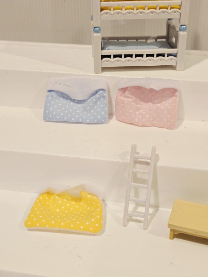 Puede incluir: Un juego de dormitorio de casa de mu&ntilde;ecas en miniatura con una litera blanca y detalles en azul y amarillo. Incluye una almohada azul y blanca con lunares, una almohada rosa y blanca con lunares, una manta amarilla y blanca con lunares, una escalera blanca y una peque&ntilde;a mesa amarilla.