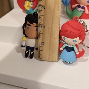 May include: Ariel and Prince Eric character keychains. The Prince is approximately 2 inches tall. Ariel is wearing a blue dress and has red hair. The Prince is wearing a white jacket with gold accents and black pants. Other Ariel keychains are visible in the background.