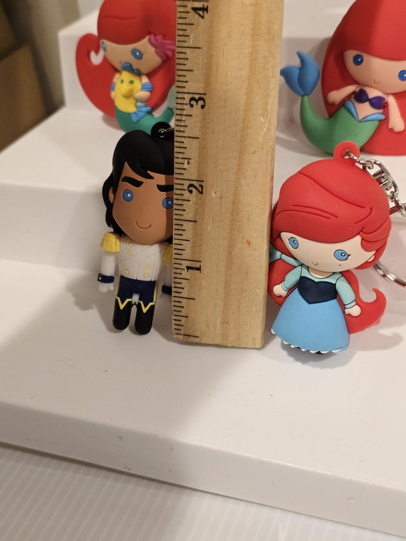 May include: Ariel and Prince Eric character keychains. The Prince is approximately 2 inches tall. Ariel is wearing a blue dress and has red hair. The Prince is wearing a white jacket with gold accents and black pants. Other Ariel keychains are visible in the background.