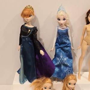 May include: A display of dolls featuring characters from an animated film. One doll is dressed in a black and blue gown with a purple skirt and a gold crown. Another doll wears a blue dress with a snowflake pattern and a silver crown. Other dolls are also visible.
