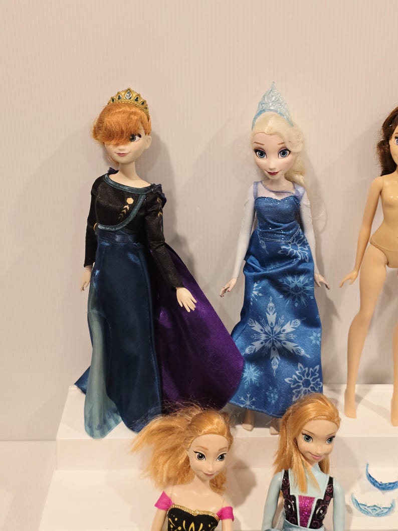 May include: A display of dolls featuring characters from an animated film. One doll is dressed in a black and blue gown with a purple skirt and a gold crown. Another doll wears a blue dress with a snowflake pattern and a silver crown. Other dolls are also visible.