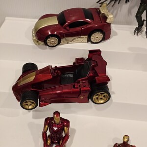 May include: A collection of toy cars and action figures. The cars are primarily red with gold accents. One car has a spoiler. The action figures include Iron Man and a villain with large wings. The toys are arranged on a white surface.