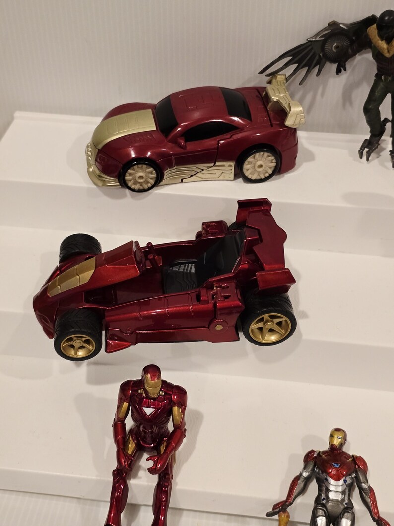 May include: A collection of toy cars and action figures. The cars are primarily red with gold accents. One car has a spoiler. The action figures include Iron Man and a villain with large wings. The toys are arranged on a white surface.