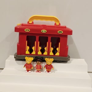 K&ouml;nnte beinhalten: Ein roter Spielzeug-Zugwagen mit gelbem Griff und drei kleinen Figuren. Der Wagen hat drei offene Fenster und eine graue Basis. Die Figuren tragen rote Jacken und Kleider. Die Figuren stehen auf einer wei&szlig;en Oberfl&auml;che.