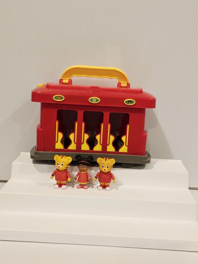 K&ouml;nnte beinhalten: Ein roter Spielzeug-Zugwagen mit gelbem Griff und drei kleinen Figuren. Der Wagen hat drei offene Fenster und eine graue Basis. Die Figuren tragen rote Jacken und Kleider. Die Figuren stehen auf einer wei&szlig;en Oberfl&auml;che.