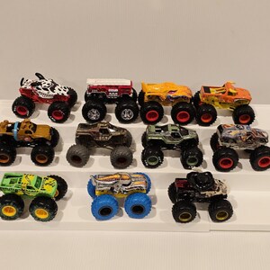 Op de afbeelding: Een verzameling van twaalf speelgoed monstertrucks in verschillende kleuren en ontwerpen. De trucks hebben grote banden en gedetailleerde carrosserie&euml;n, waaronder een truck met Dalmatische thema, een rode truck met een wit dak en een groene truck met gele accenten.