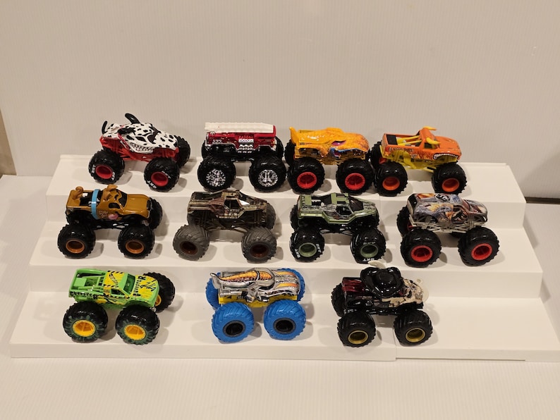 Op de afbeelding: Een verzameling van twaalf speelgoed monstertrucks in verschillende kleuren en ontwerpen. De trucks hebben grote banden en gedetailleerde carrosserie&euml;n, waaronder een truck met Dalmatische thema, een rode truck met een wit dak en een groene truck met gele accenten.