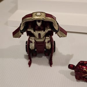 May include: A red and gold toy robot with a detailed design. The robot has a helmet and chest plate with gold accents. The toy is on a white surface.