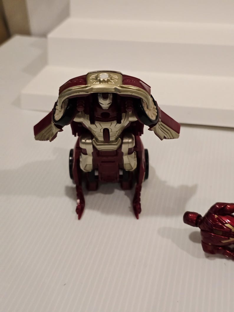 May include: A red and gold toy robot with a detailed design. The robot has a helmet and chest plate with gold accents. The toy is on a white surface.