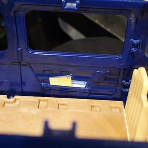 May include: Interior view of a blue toy vehicle, featuring a door with a window and a handle. A small compartment holds a yellow card labeled "Map" and a white card labeled "Village Pack". The interior has tan seating.