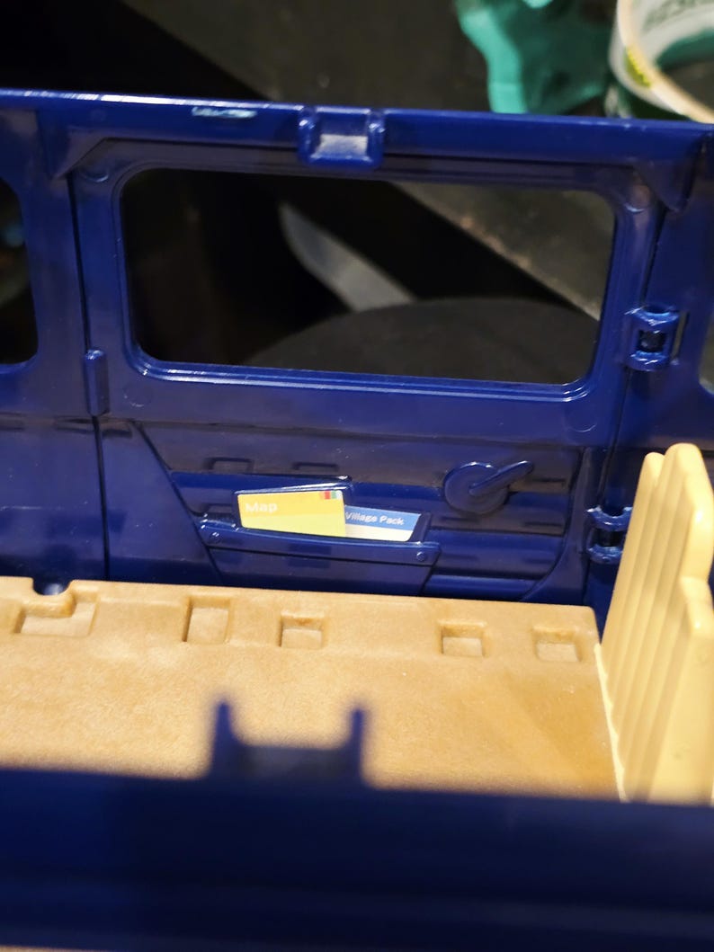 May include: Interior view of a blue toy vehicle, featuring a door with a window and a handle. A small compartment holds a yellow card labeled "Map" and a white card labeled "Village Pack". The interior has tan seating.