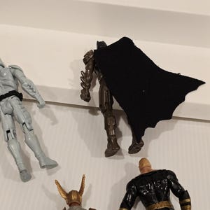 May include: A collection of superhero action figures, including Batman with a black cape, a gray armored figure, a figure with a golden helmet, and a black and gold figure. The figures are on a white surface.