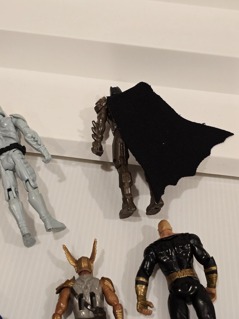 May include: A collection of superhero action figures, including Batman with a black cape, a gray armored figure, a figure with a golden helmet, and a black and gold figure. The figures are on a white surface.