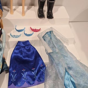 May include: A selection of doll accessories, including a blue cape with floral embroidery, a light blue dress, and several tiaras. Doll legs and black boots are also visible.