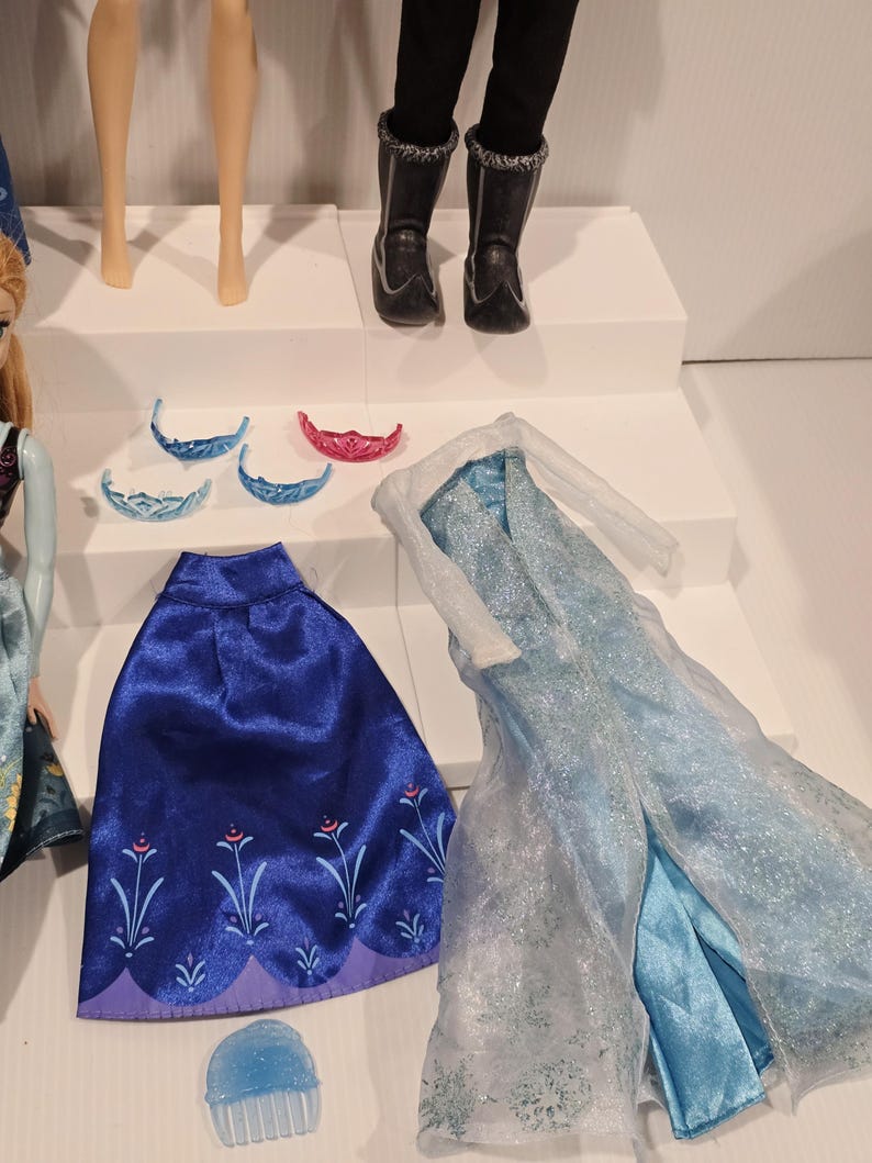 May include: A selection of doll accessories, including a blue cape with floral embroidery, a light blue dress, and several tiaras. Doll legs and black boots are also visible.