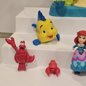 May include: A collection of colorful Little Mermaid character figurines. Includes a yellow and blue fish, a red crab, a small red crab, and a figurine of Ariel in a blue and pink dress.