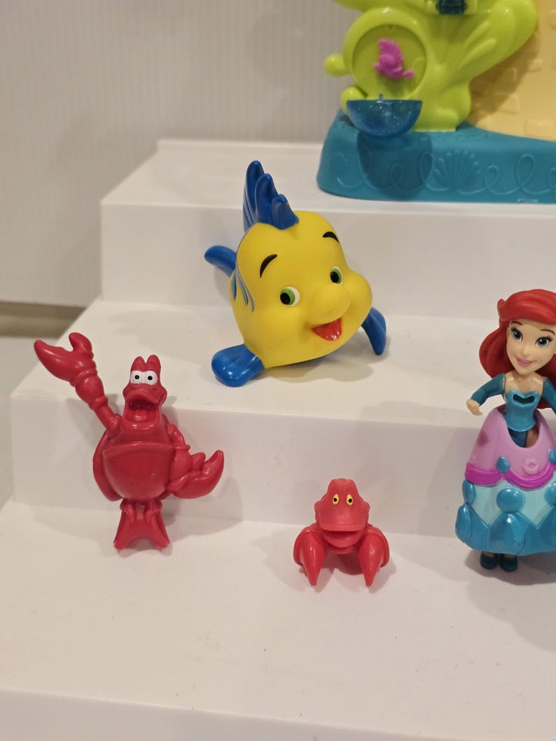 May include: A collection of colorful Little Mermaid character figurines. Includes a yellow and blue fish, a red crab, a small red crab, and a figurine of Ariel in a blue and pink dress.