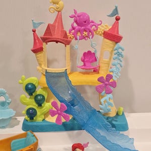 K&ouml;nnte beinhalten: Ein farbenfrohes Kunststoff-Spielset mit einem Schloss mit rosa und gelben T&uuml;rmen, einer blauen Rutsche und Dekorationen zum Thema Meer. Enth&auml;lt eine kleine Ariel-Figur und anderes Spielzeug mit Meeresthema. Das Set ist f&uuml;r Kinder konzipiert.