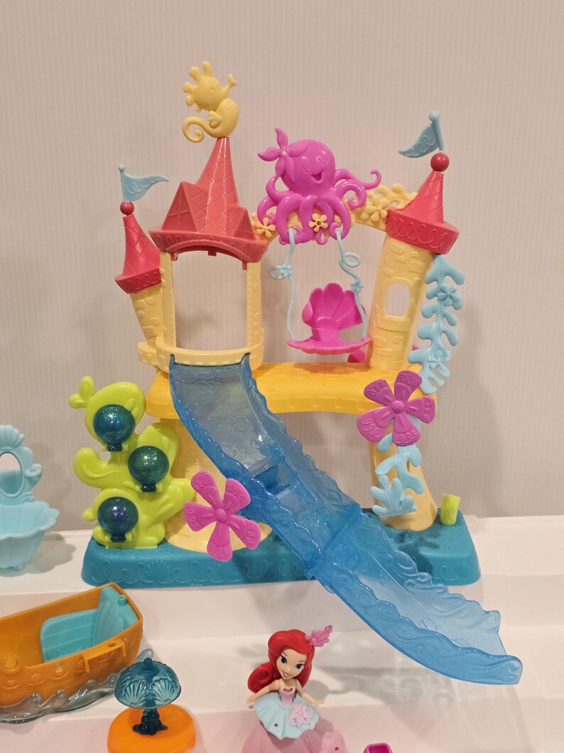 K&ouml;nnte beinhalten: Ein farbenfrohes Kunststoff-Spielset mit einem Schloss mit rosa und gelben T&uuml;rmen, einer blauen Rutsche und Dekorationen zum Thema Meer. Enth&auml;lt eine kleine Ariel-Figur und anderes Spielzeug mit Meeresthema. Das Set ist f&uuml;r Kinder konzipiert.