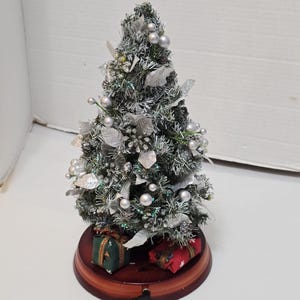 May include: A miniature Christmas tree with silver ornaments and decorative leaves. The tree sits on a round, wooden base with two small gift boxes. A black power cord extends from the base, suggesting it may light up.