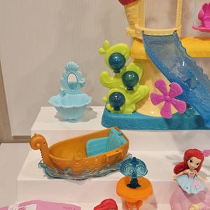 K&ouml;nnte beinhalten: Ariel's Unterwasser-Schloss-Spielset mit einem gelb-rosa Schloss, einer blauen Rutsche, einem Boot und Figuren von Arielle und Sebastian. Enth&auml;lt einen blauen Schminktisch, einen orangen Tisch mit einer Lampe und eine rote Krabbe.