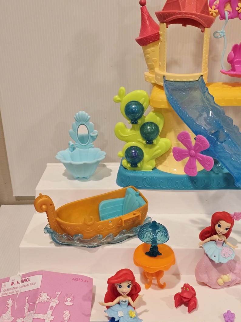 K&ouml;nnte beinhalten: Ariel's Unterwasser-Schloss-Spielset mit einem gelb-rosa Schloss, einer blauen Rutsche, einem Boot und Figuren von Arielle und Sebastian. Enth&auml;lt einen blauen Schminktisch, einen orangen Tisch mit einer Lampe und eine rote Krabbe.
