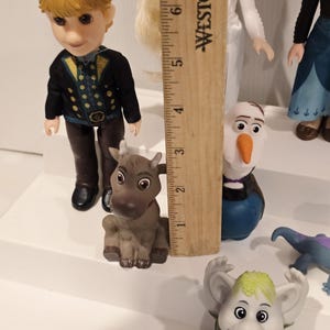 May include: A collection of Frozen character figurines, including Kristoff, Olaf, and a troll. Kristoff is wearing a black jacket with gold accents. The figurines are on a white surface. A wooden ruler is in the background.