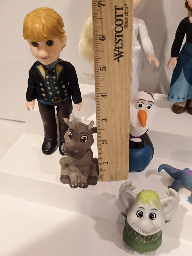 May include: A collection of Frozen character figurines, including Kristoff, Olaf, and a troll. Kristoff is wearing a black jacket with gold accents. The figurines are on a white surface. A wooden ruler is in the background.