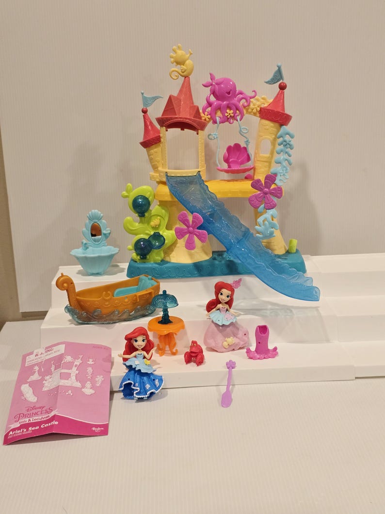 K&ouml;nnte beinhalten: Ariel's Sea Castle Spielset mit einem farbenfrohen Schloss, einer Rutsche, einem Boot und Figuren. Das Schloss hat rosa, gelbe und blaue Akzente, mit einer Rutsche und dekorativen Elementen. Enth&auml;lt zwei Ariel-Figuren, ein Boot und Zubeh&ouml;r.